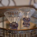 Two clear faceted cocktail glasses with purple bases, displayed on a decorative mirrored tray with a patterned metal rim.