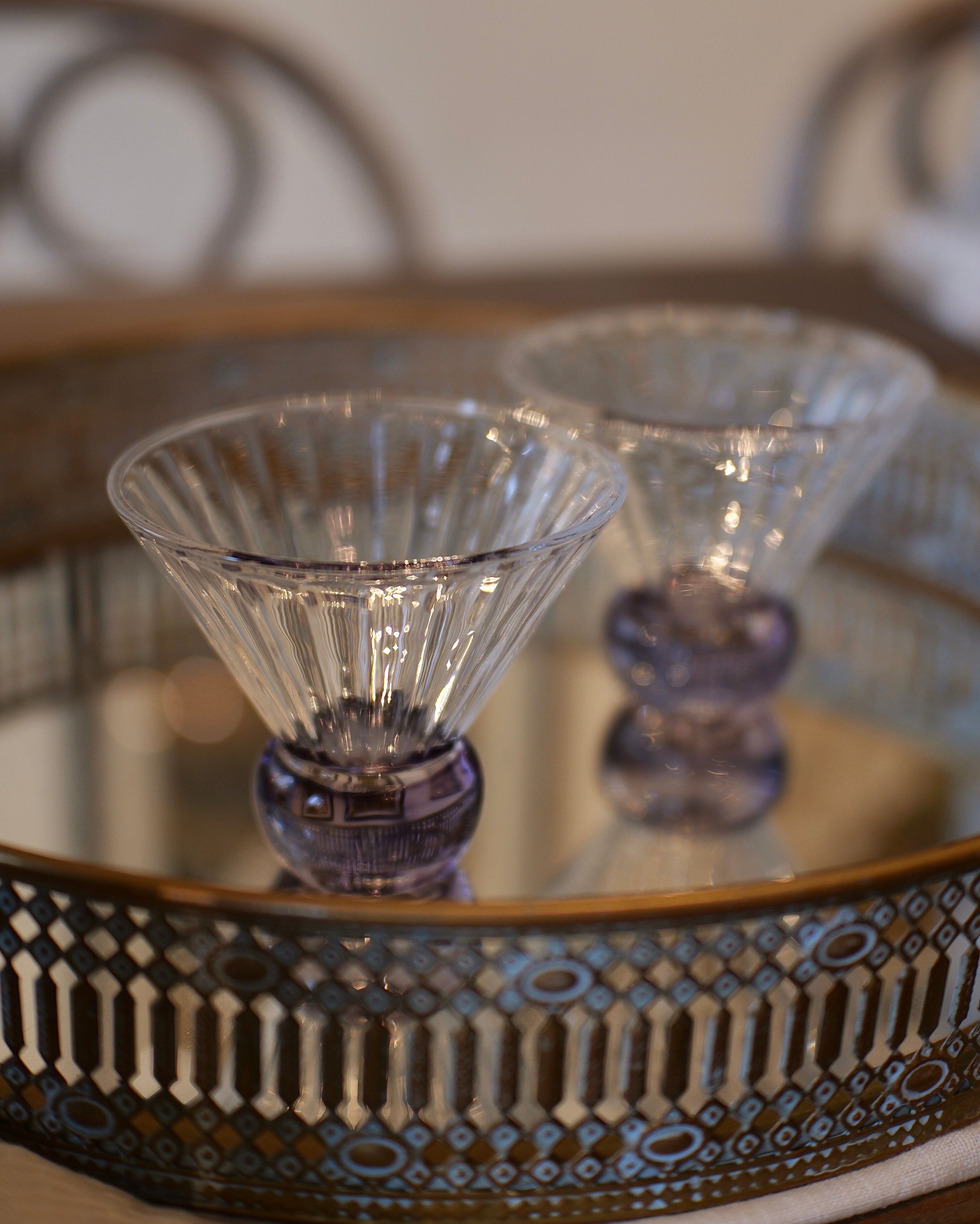 Two clear faceted cocktail glasses with purple bases, displayed on a decorative mirrored tray with a patterned metal rim.