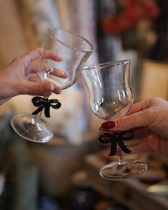 Two hands holding clear stemmed wine glasses with small black ribbon bows at the base, clinking together in a toast. The background is a softly blurred indoor setting.