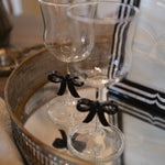 Two clear stemmed drinking glasses with black ribbon bows tied near the stems, displayed on a mirrored tray in an indoor setting.