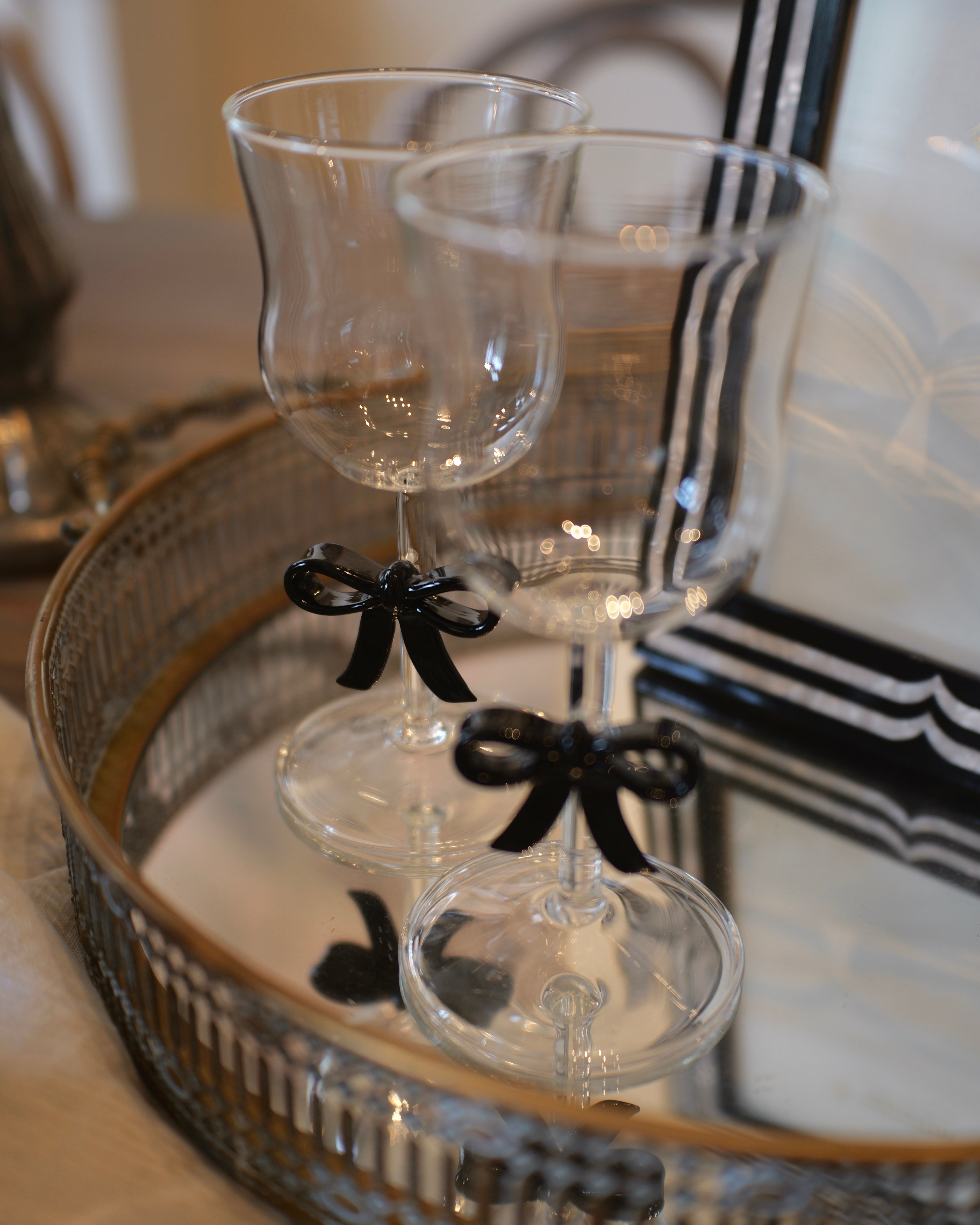Two clear stemmed drinking glasses with black ribbon bows tied near the stems, displayed on a mirrored tray in an indoor setting.
