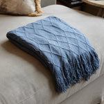 A folded slate blue knit throw blanket with a diamond textured pattern and fringed edges, resting on a light beige upholstered ottoman in a cozy indoor setting.