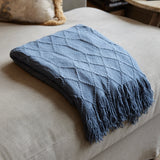 A folded slate blue knit throw blanket with a diamond textured pattern and fringed edges, resting on a light beige upholstered ottoman in a cozy indoor setting.