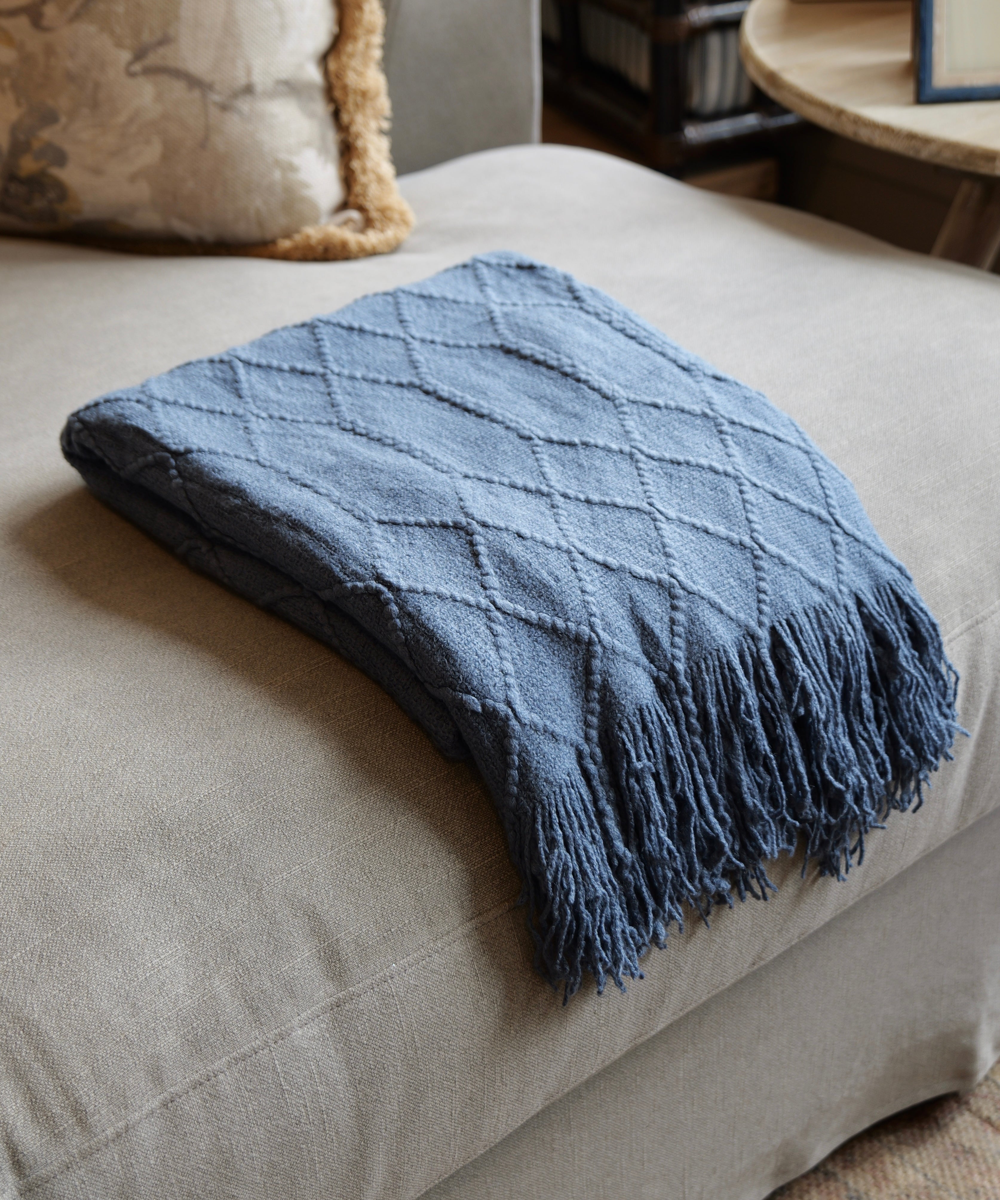 A folded slate blue knit throw blanket with a diamond textured pattern and fringed edges, resting on a light beige upholstered ottoman in a cozy indoor setting.