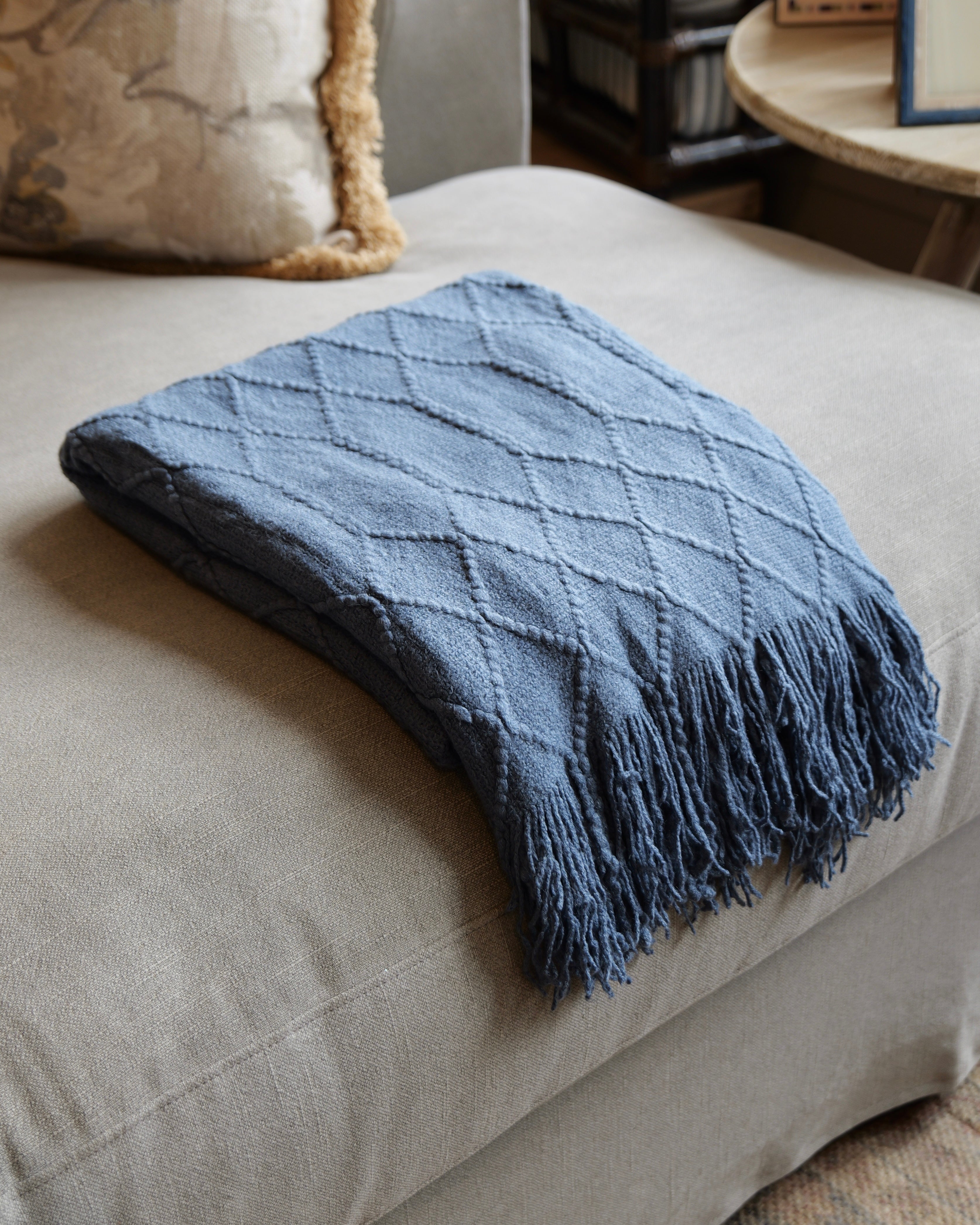 A folded slate blue knit throw blanket with a diamond textured pattern and fringed edges, resting on a light beige upholstered ottoman in a cozy indoor setting.