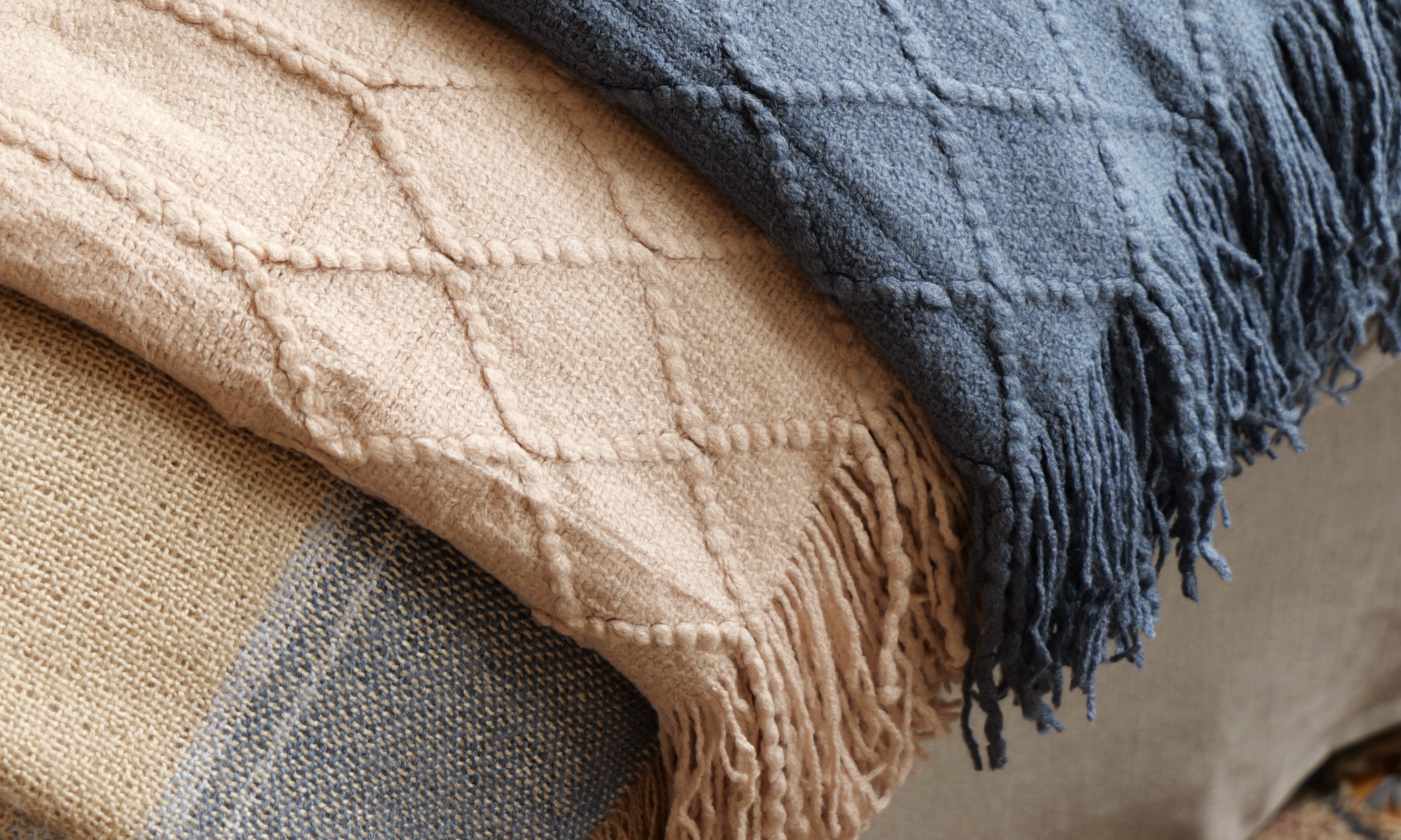 A stack of three textured woven throw blankets in navy blue, beige, and tan, draped over a sofa in a cozy indoor setting.