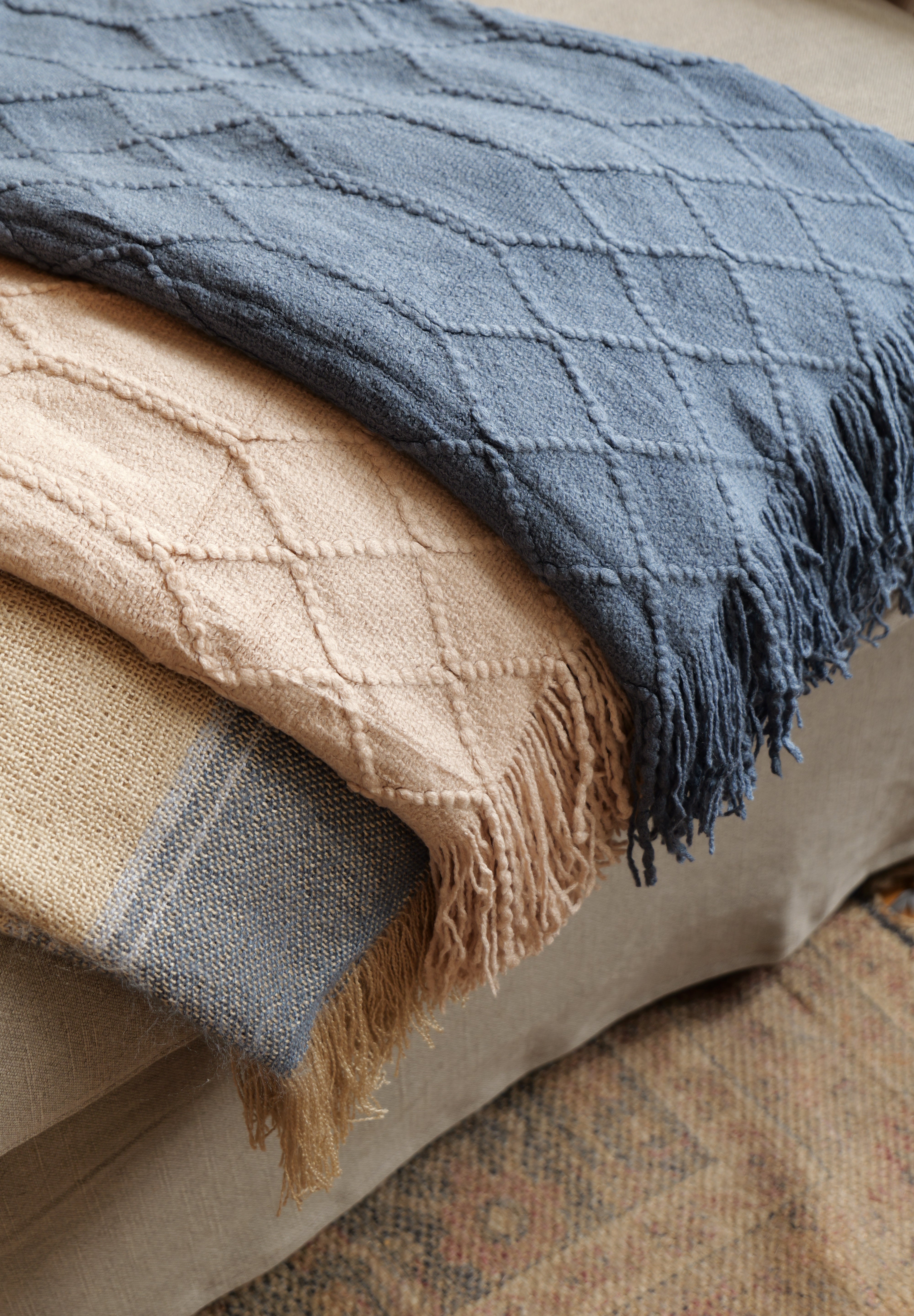 A stack of three textured woven throw blankets in navy blue, beige, and tan, draped over a sofa in a cozy indoor setting.
