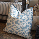 A square throw pillow with a blue floral vine pattern on a cream background, styled on a patterned rug in front of a striped sofa.