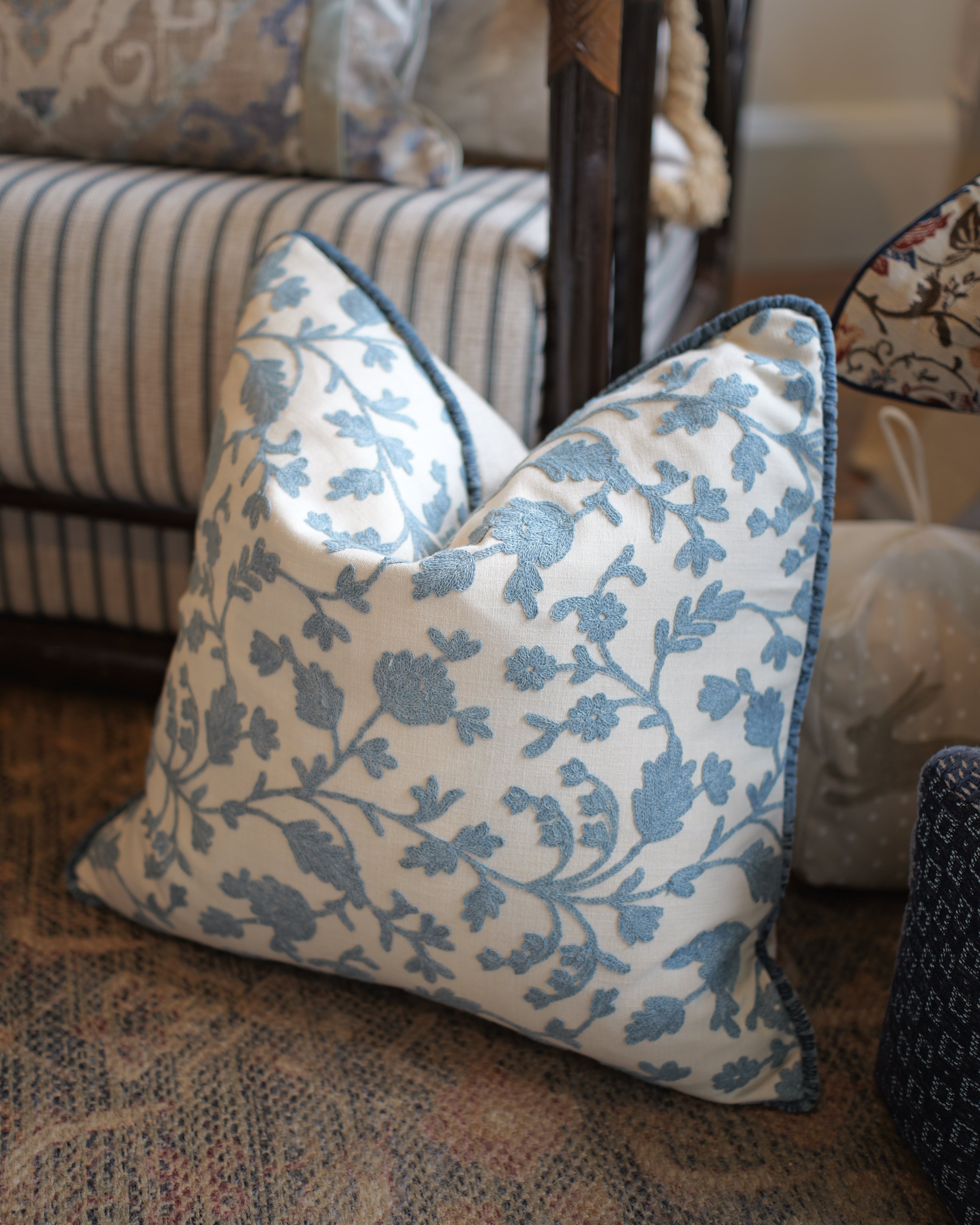 A square throw pillow with a blue floral vine pattern on a cream background, styled on a patterned rug in front of a striped sofa.