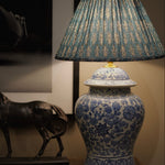 A blue and white porcelain-style table lamp with a pleated blue and beige patterned shade, displayed on a wooden surface next to a dark horse figurine.
