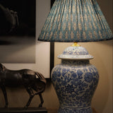 A blue and white porcelain-style table lamp with a pleated blue and beige patterned shade, displayed on a wooden surface next to a dark horse figurine.