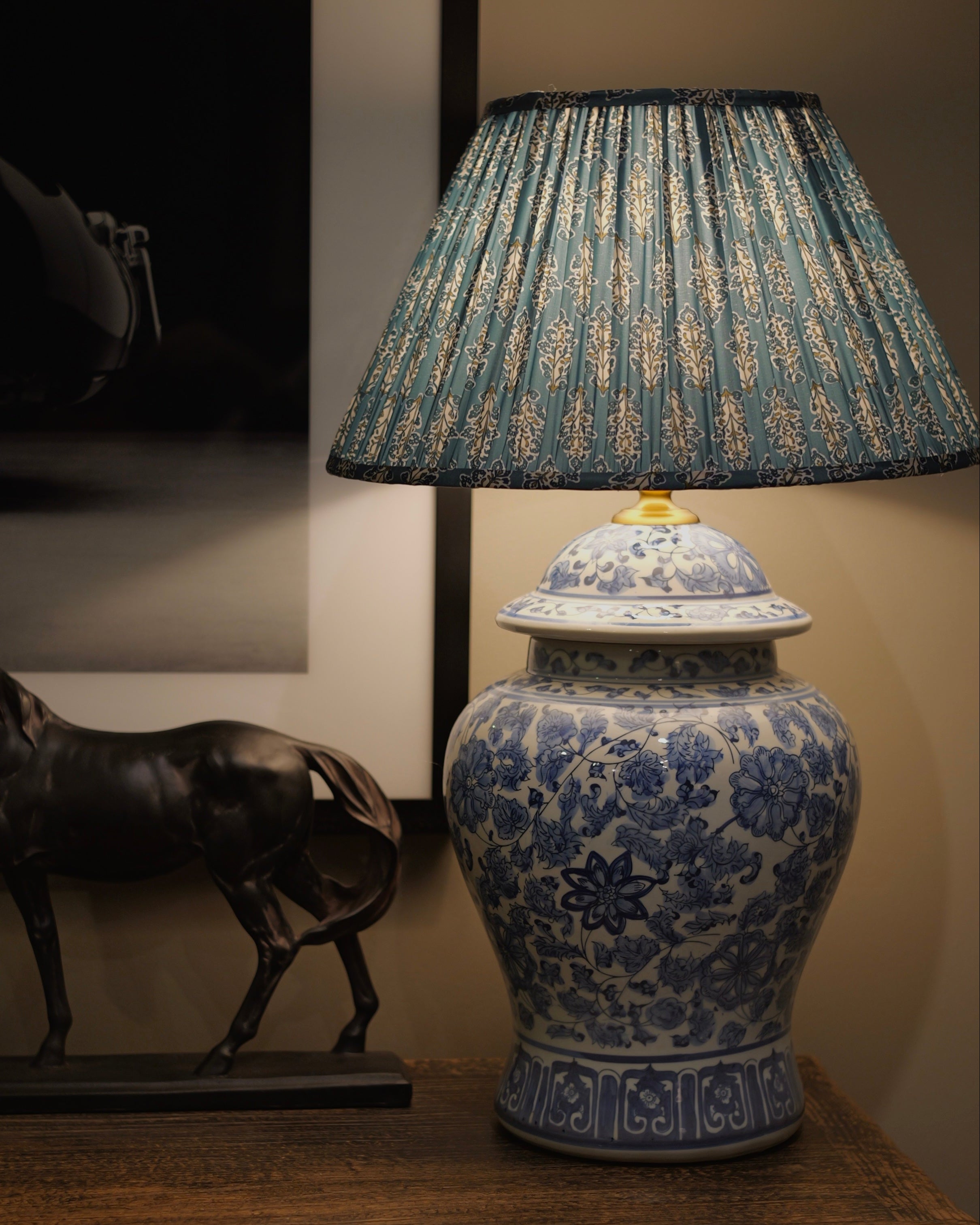 A blue and white porcelain-style table lamp with a pleated blue and beige patterned shade, displayed on a wooden surface next to a dark horse figurine.