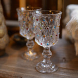Two clear glass goblets with colorful faceted jewel accents, displayed on a rustic wooden surface in a warm, softly blurred setting.