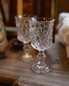 Two clear glass goblets with colorful faceted jewel accents, displayed on a rustic wooden surface in a warm, softly blurred setting.
