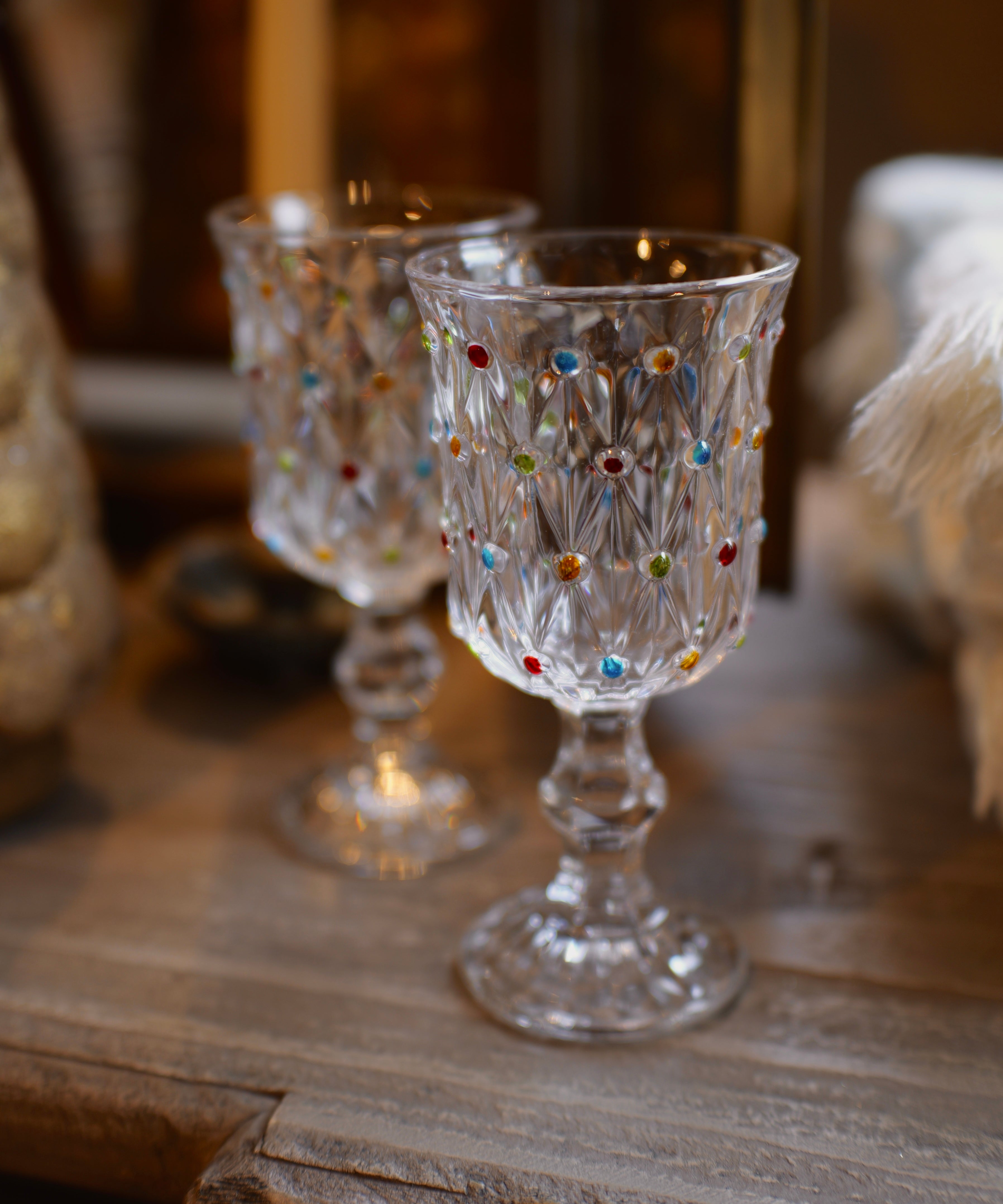 Two clear glass goblets with colorful faceted jewel accents, displayed on a rustic wooden surface in a warm, softly blurred setting.