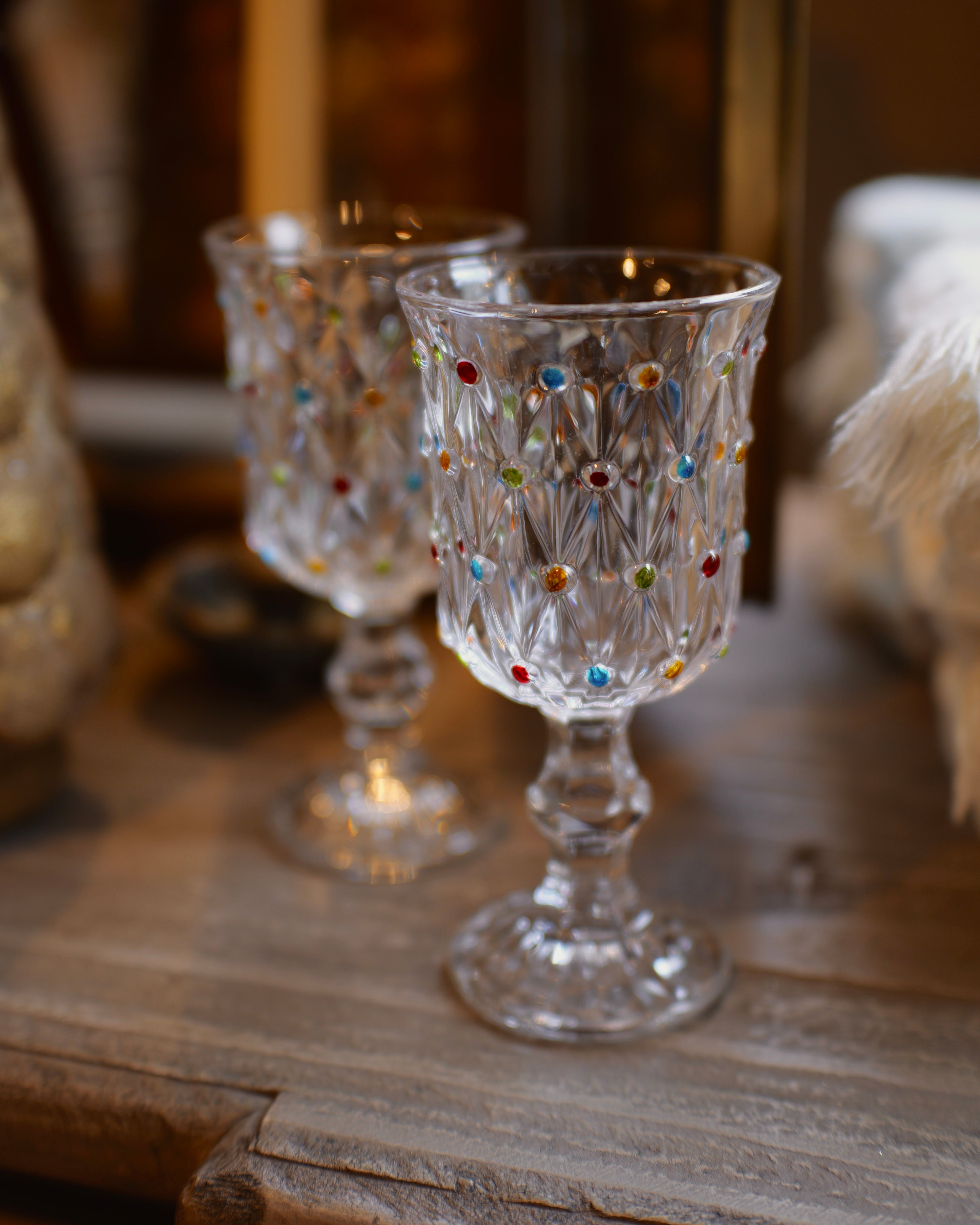 Two clear glass goblets with colorful faceted jewel accents, displayed on a rustic wooden surface in a warm, softly blurred setting.