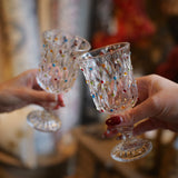 Two hands holding clear cut-crystal goblets with colorful rhinestone accents, clinking together in a toast. The background is a softly blurred shop interior.