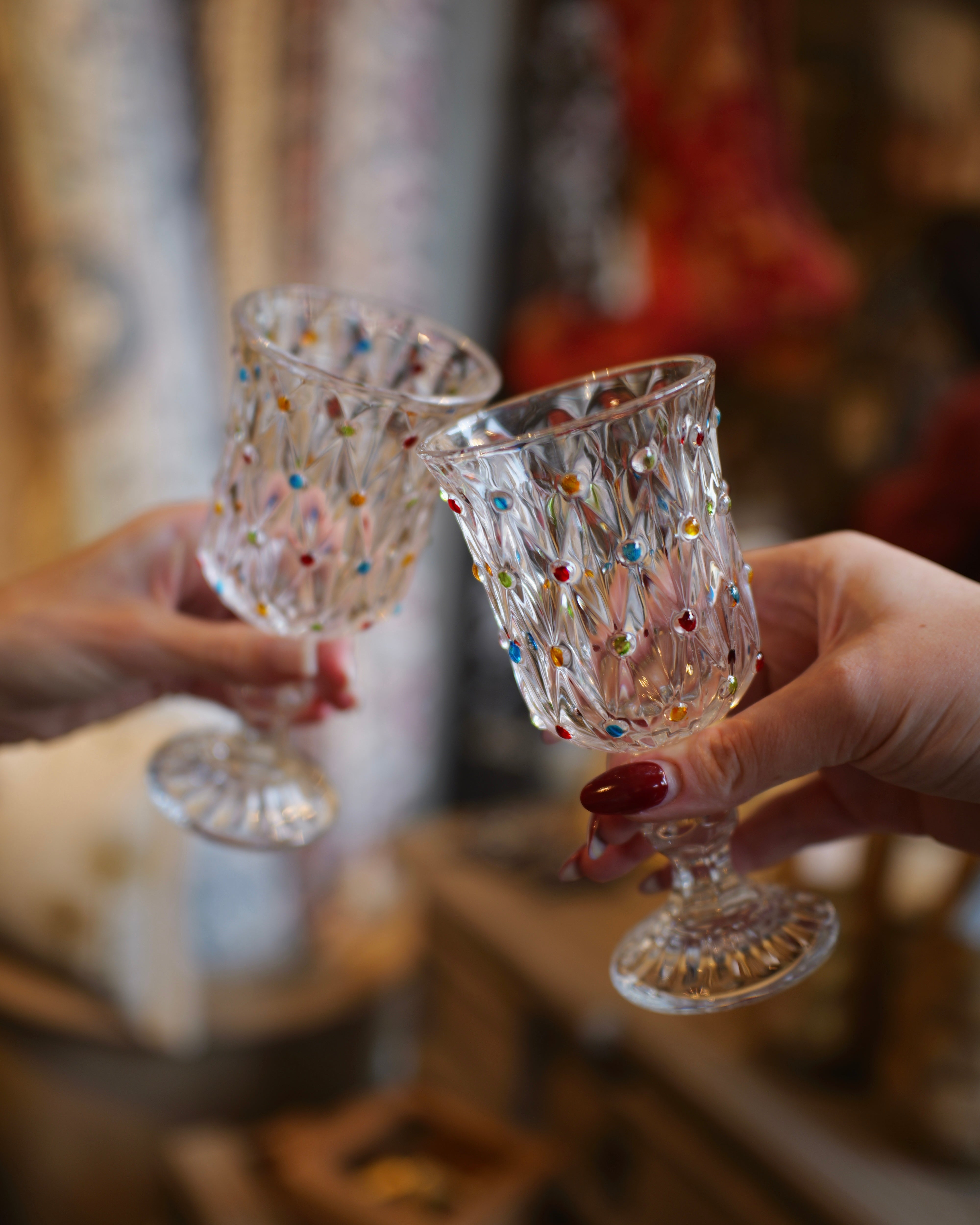 Two hands holding clear cut-crystal goblets with colorful rhinestone accents, clinking together in a toast. The background is a softly blurred shop interior.