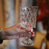 A hand holding a clear cut-glass goblet decorated with small multicolored heart-shaped accents, set against a blurred warm-toned background.