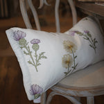 A white rectangular throw pillow with embroidered purple and yellow thistle flowers, resting on a distressed white wooden chair in a rustic indoor setting.
