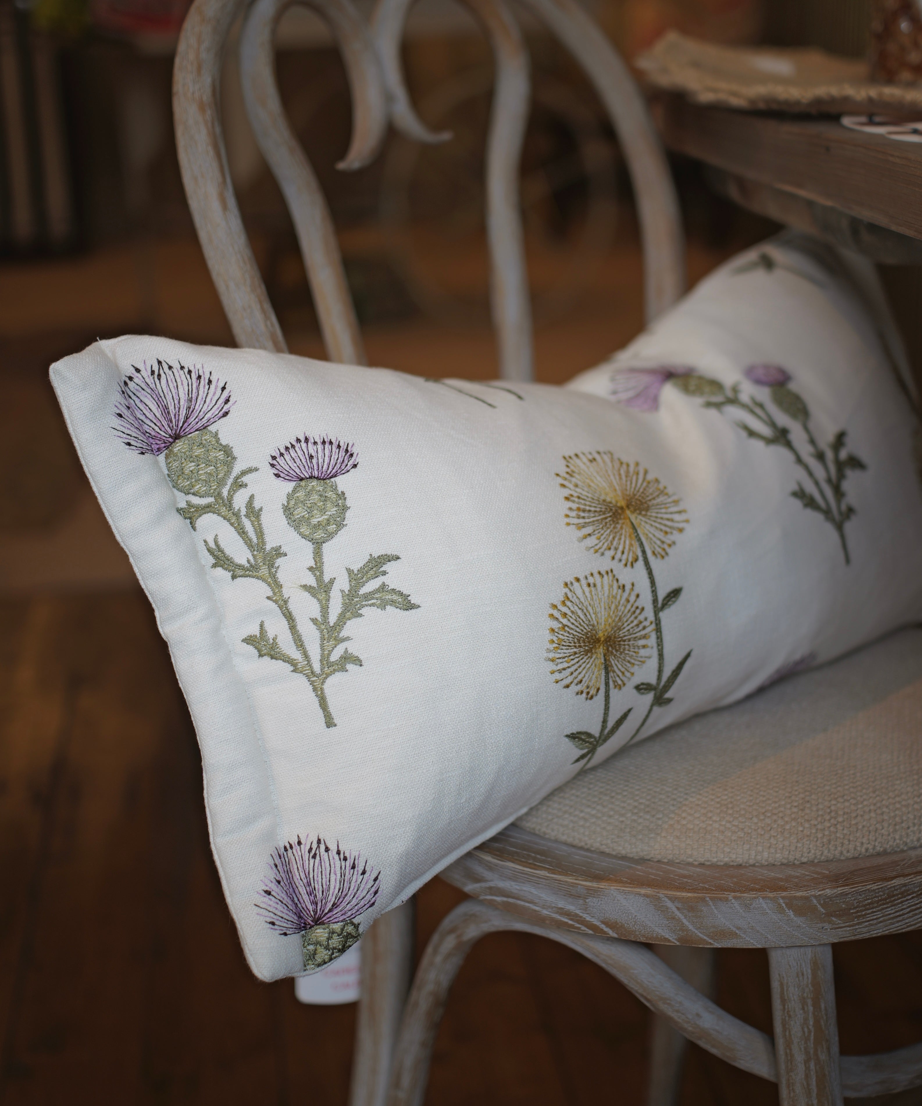 A white rectangular throw pillow with embroidered purple and yellow thistle flowers, resting on a distressed white wooden chair in a rustic indoor setting.