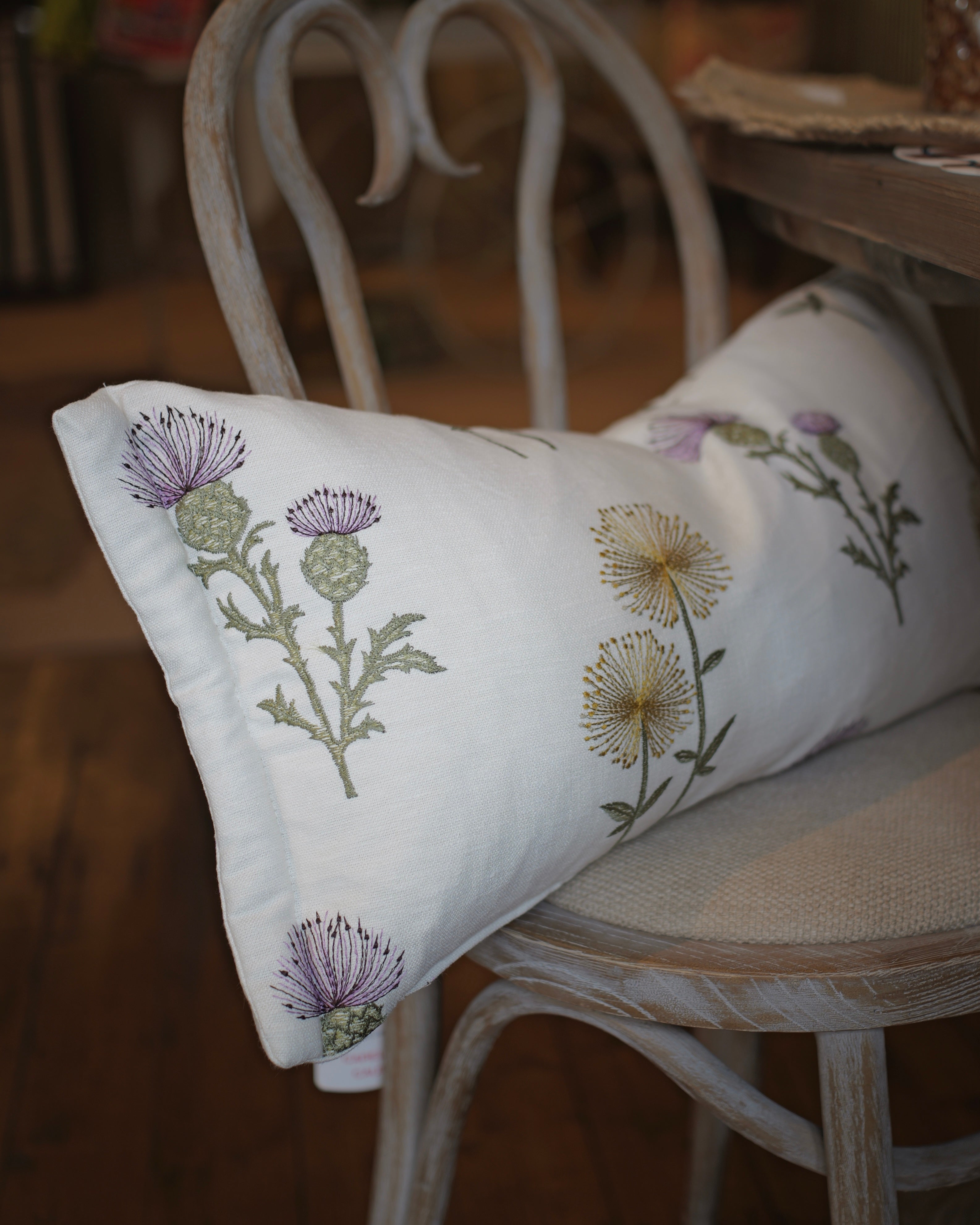 A white rectangular throw pillow with embroidered purple and yellow thistle flowers, resting on a distressed white wooden chair in a rustic indoor setting.