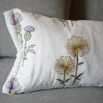 A rectangular white throw pillow with embroidered purple thistles and yellow dandelions, resting on a gray fabric sofa.