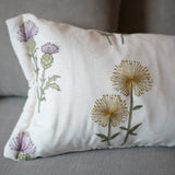A rectangular white throw pillow with embroidered purple thistles and yellow dandelions, resting on a gray fabric sofa.