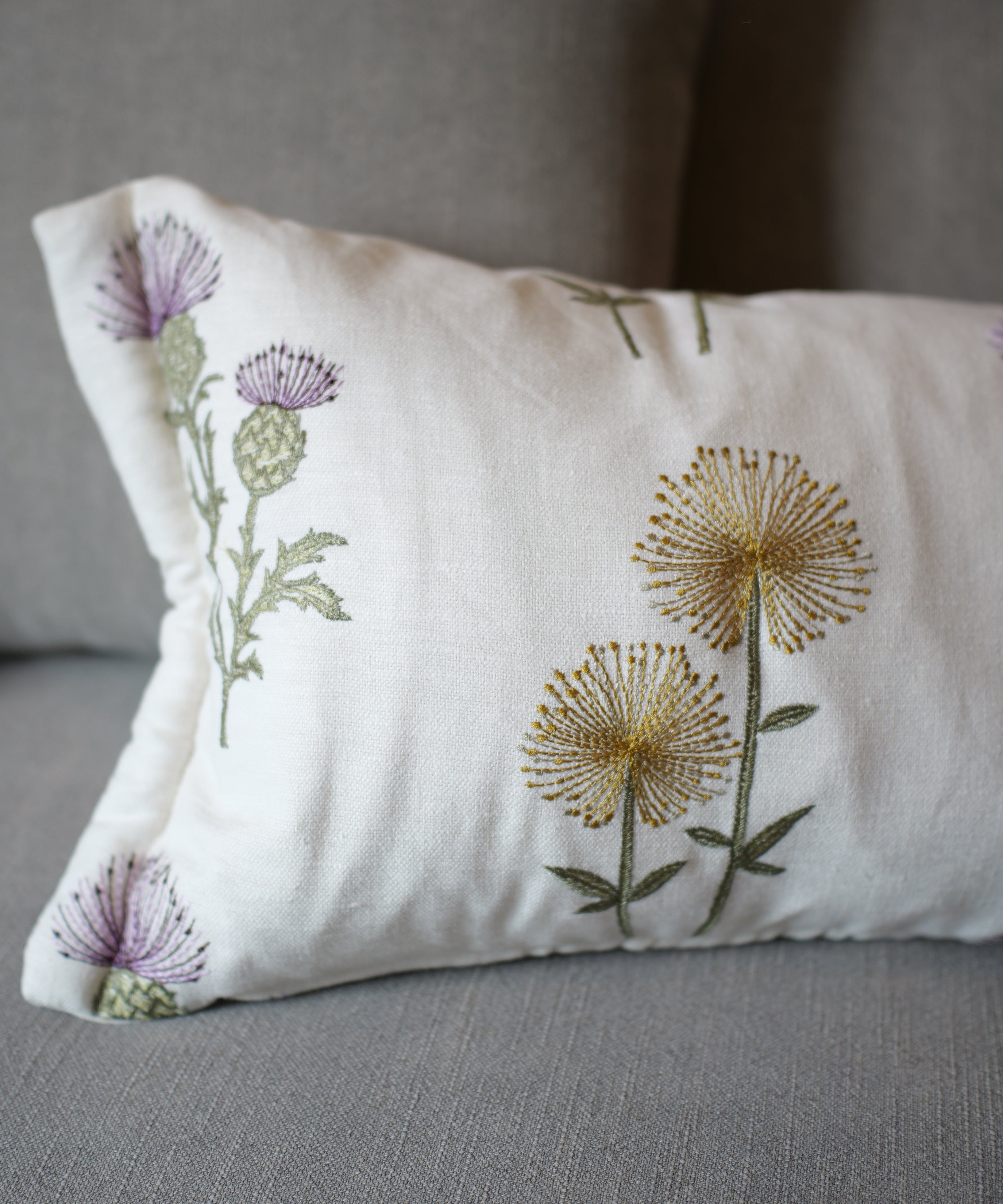 A rectangular white throw pillow with embroidered purple thistles and yellow dandelions, resting on a gray fabric sofa.