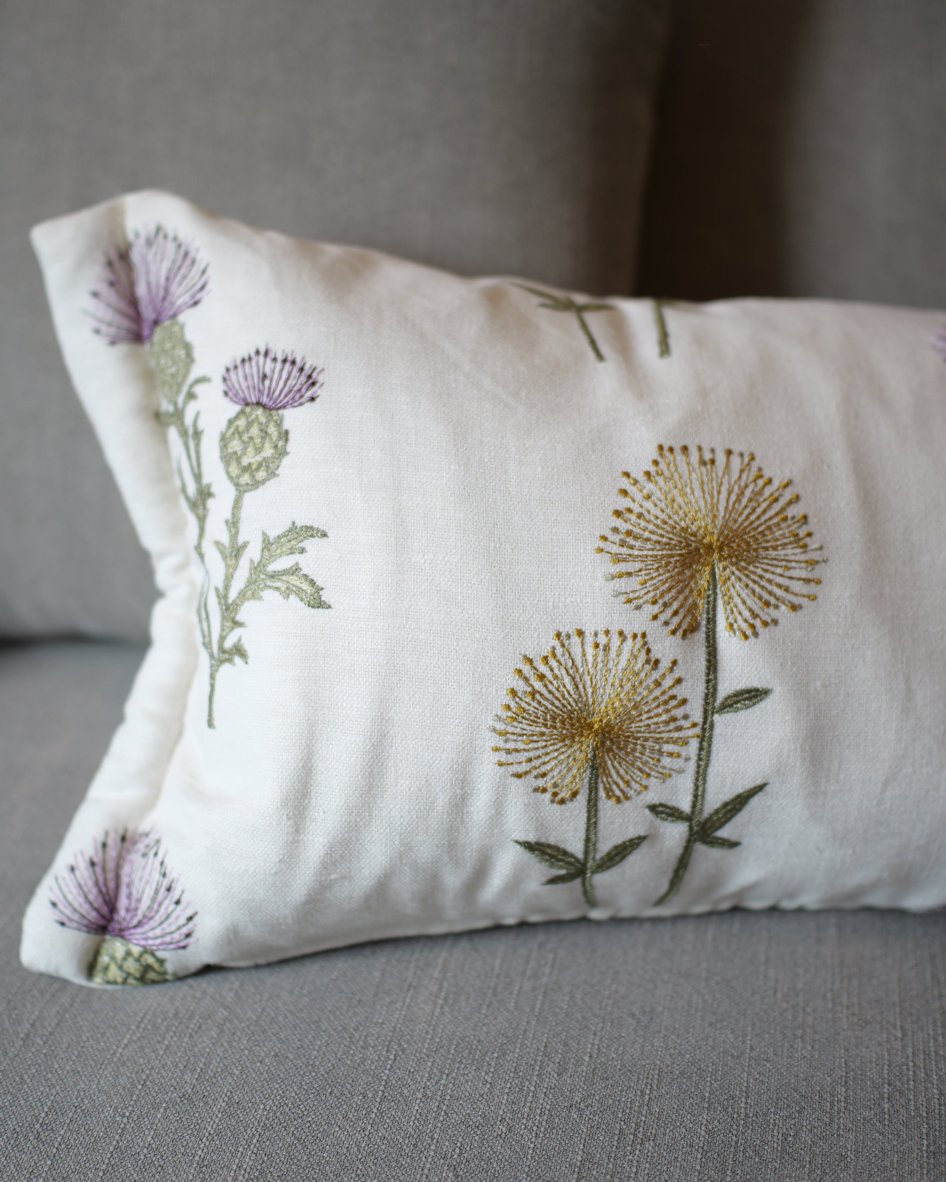 A rectangular white throw pillow with embroidered purple thistles and yellow dandelions, resting on a gray fabric sofa.