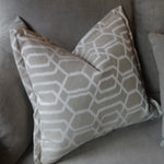 A square throw pillow with a white geometric trellis pattern on a taupe-beige background, resting on a matching gray fabric sofa.