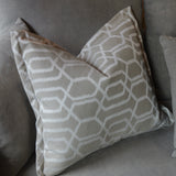 A square throw pillow with a white geometric trellis pattern on a taupe-beige background, resting on a matching gray fabric sofa.
