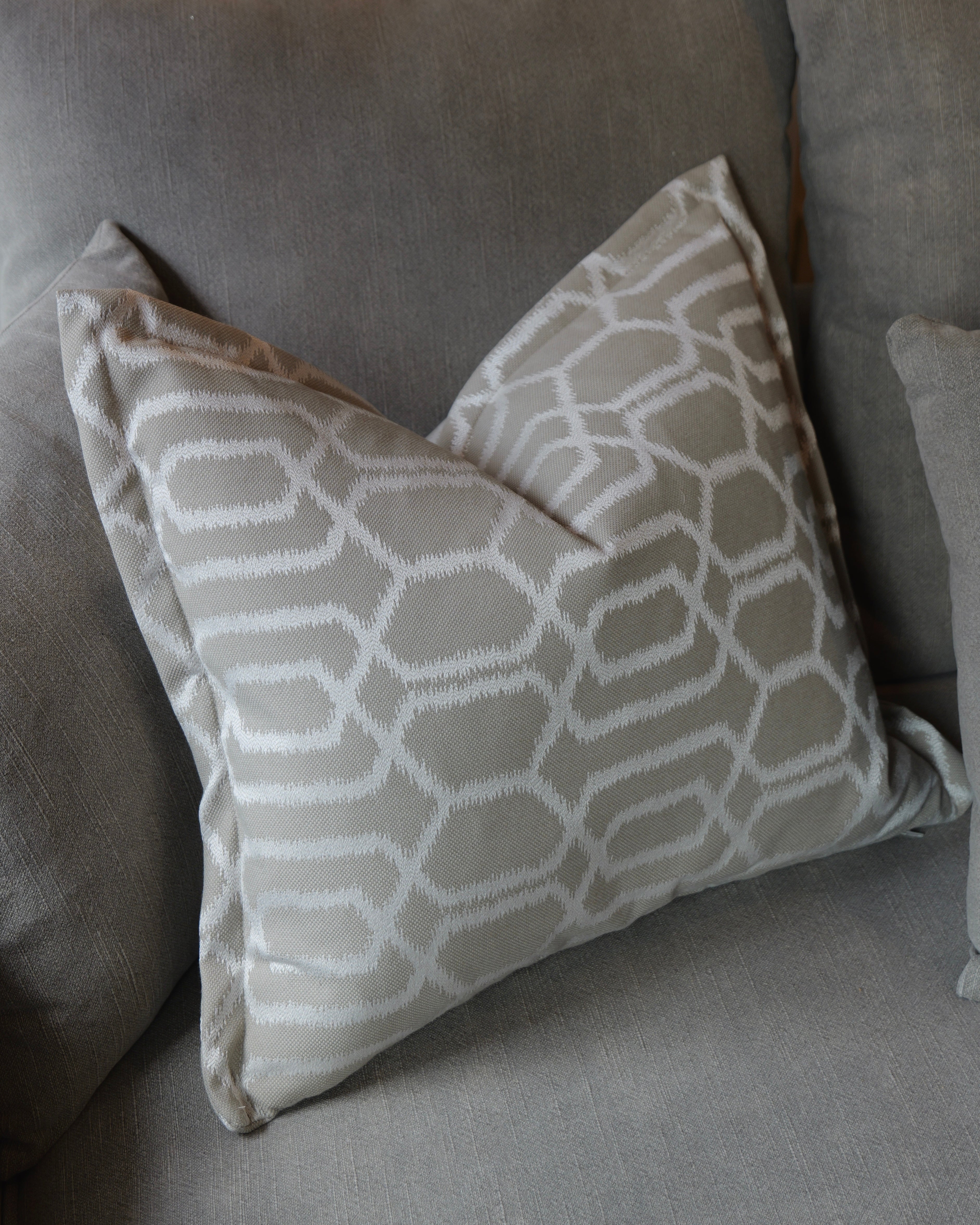A square throw pillow with a white geometric trellis pattern on a taupe-beige background, resting on a matching gray fabric sofa.