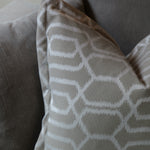 A close-up of a decorative throw pillow with a white geometric lattice pattern on a taupe-beige background, resting on a matching gray fabric sofa.