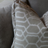 A close-up of a decorative throw pillow with a white geometric lattice pattern on a taupe-beige background, resting on a matching gray fabric sofa.
