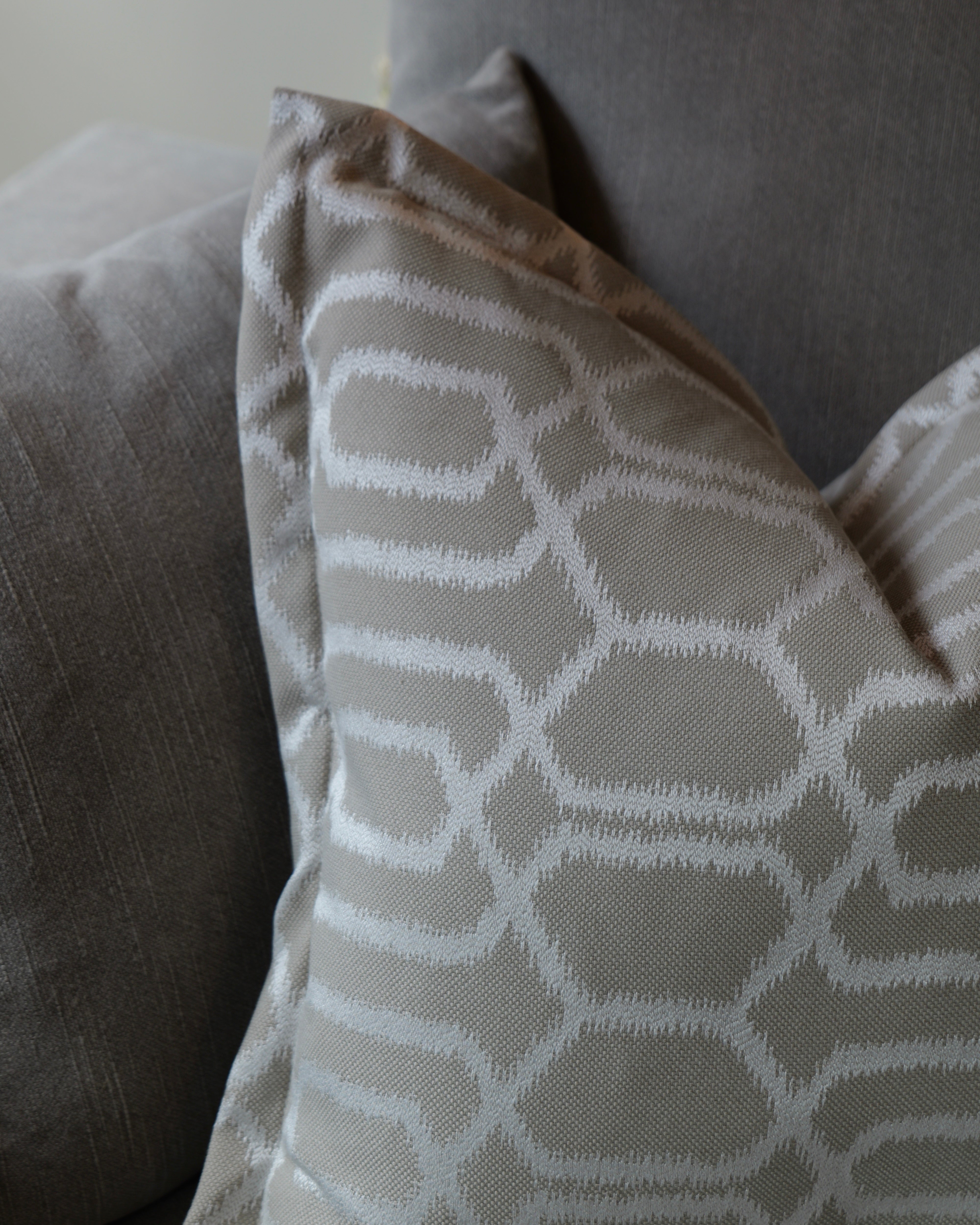 A close-up of a decorative throw pillow with a white geometric lattice pattern on a taupe-beige background, resting on a matching gray fabric sofa.