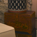 Decorative lamp on a wooden chest against a stone wall with a chair in the foreground.