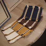 Four woven fabric swatches with fringe details, featuring navy blue, mustard yellow, and brown colours with cream and dark blue striped patterns, displayed on a light wood table.
