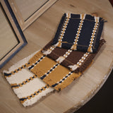 Four woven fabric swatches with fringe details, featuring navy blue, mustard yellow, and brown colours with cream and dark blue striped patterns, displayed on a light wood table.