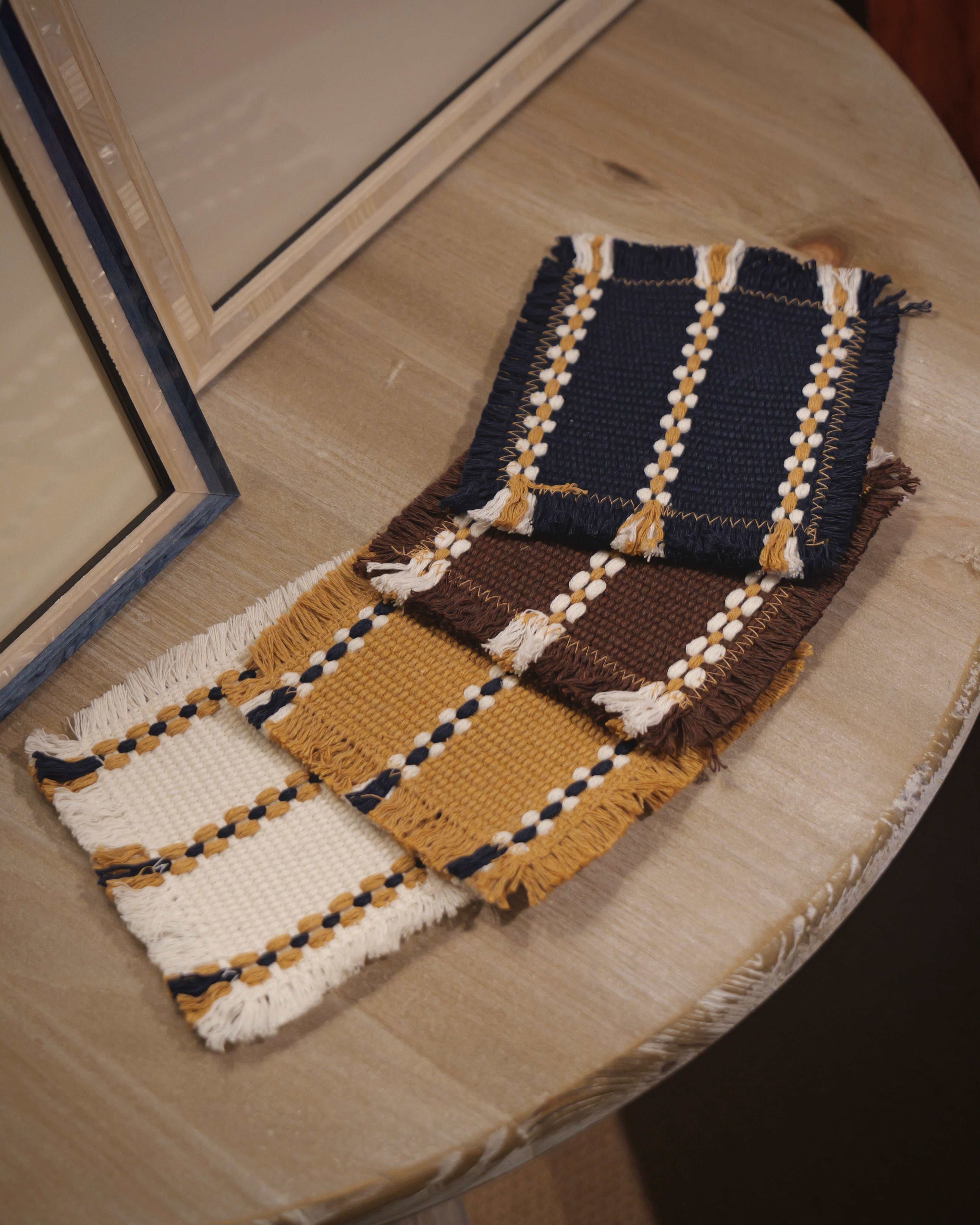 Four woven fabric swatches with fringe details, featuring navy blue, mustard yellow, and brown colours with cream and dark blue striped patterns, displayed on a light wood table.