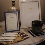 Two decorative picture frames on a rustic wooden table, one with a blue frame and one with a white frame, alongside a dark ceramic mug on a woven coaster and matching coasters.