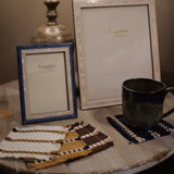 Two decorative picture frames on a rustic wooden table, one with a blue frame and one with a white frame, alongside a dark ceramic mug on a woven coaster and matching coasters.