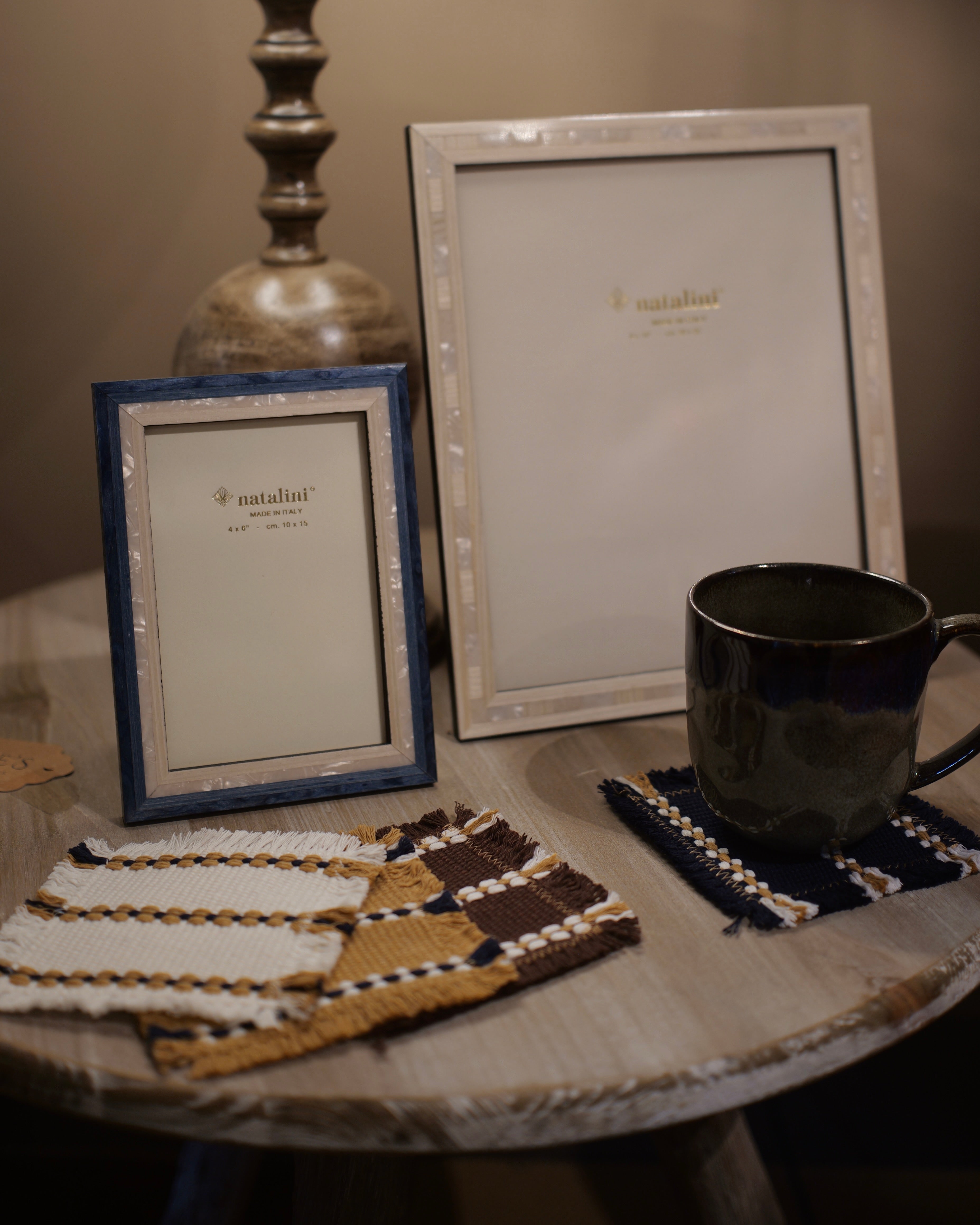 Two decorative picture frames on a rustic wooden table, one with a blue frame and one with a white frame, alongside a dark ceramic mug on a woven coaster and matching coasters.