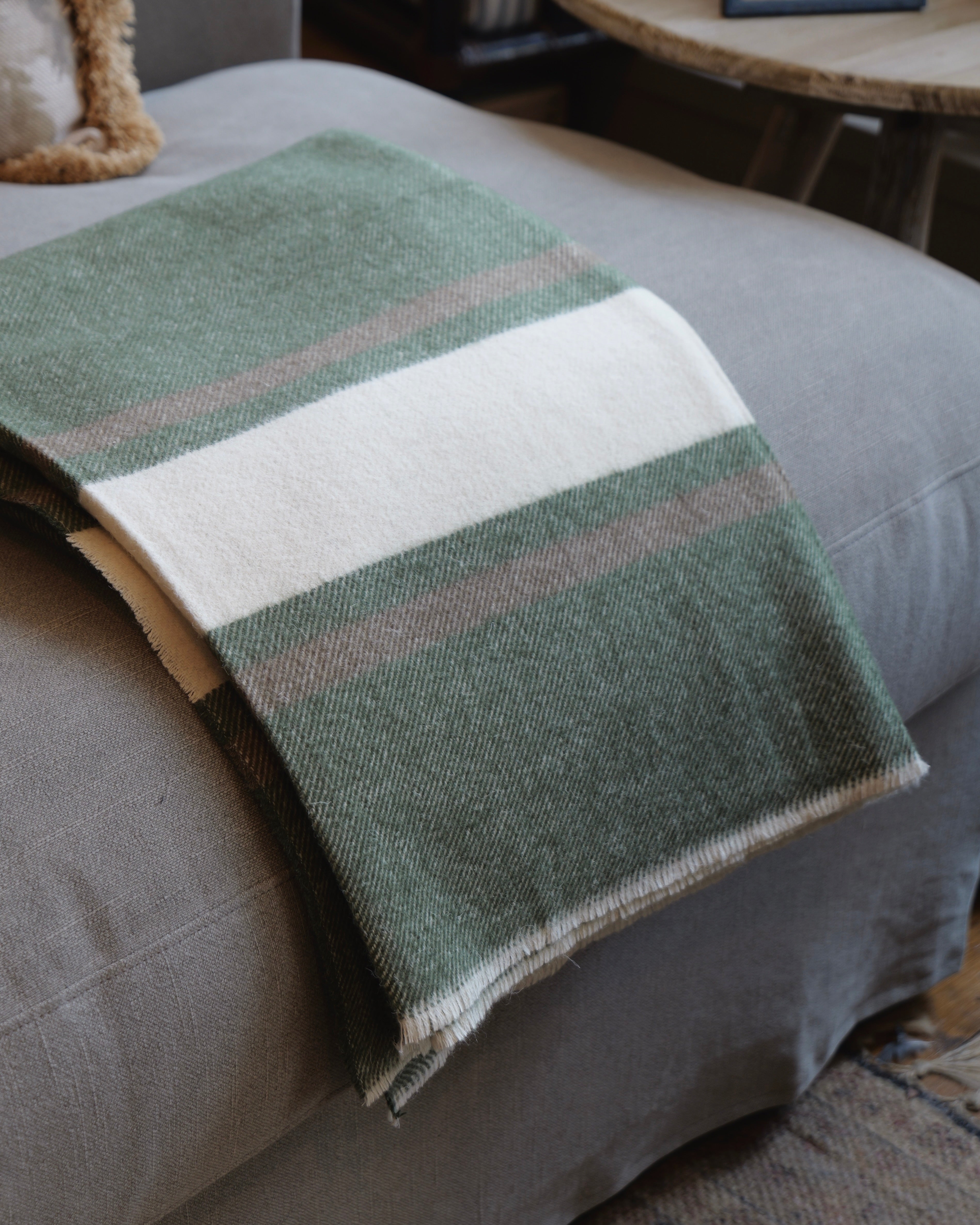 A woven throw blanket with horizontal stripes in sage green, cream, and taupe, draped over a gray upholstered ottoman in a cozy indoor setting.