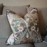 A square decorative throw pillow with a detailed botanical and floral damask pattern in muted green, beige, orange, and white, featuring a dark green piped edge, resting on a gray fabric sofa.