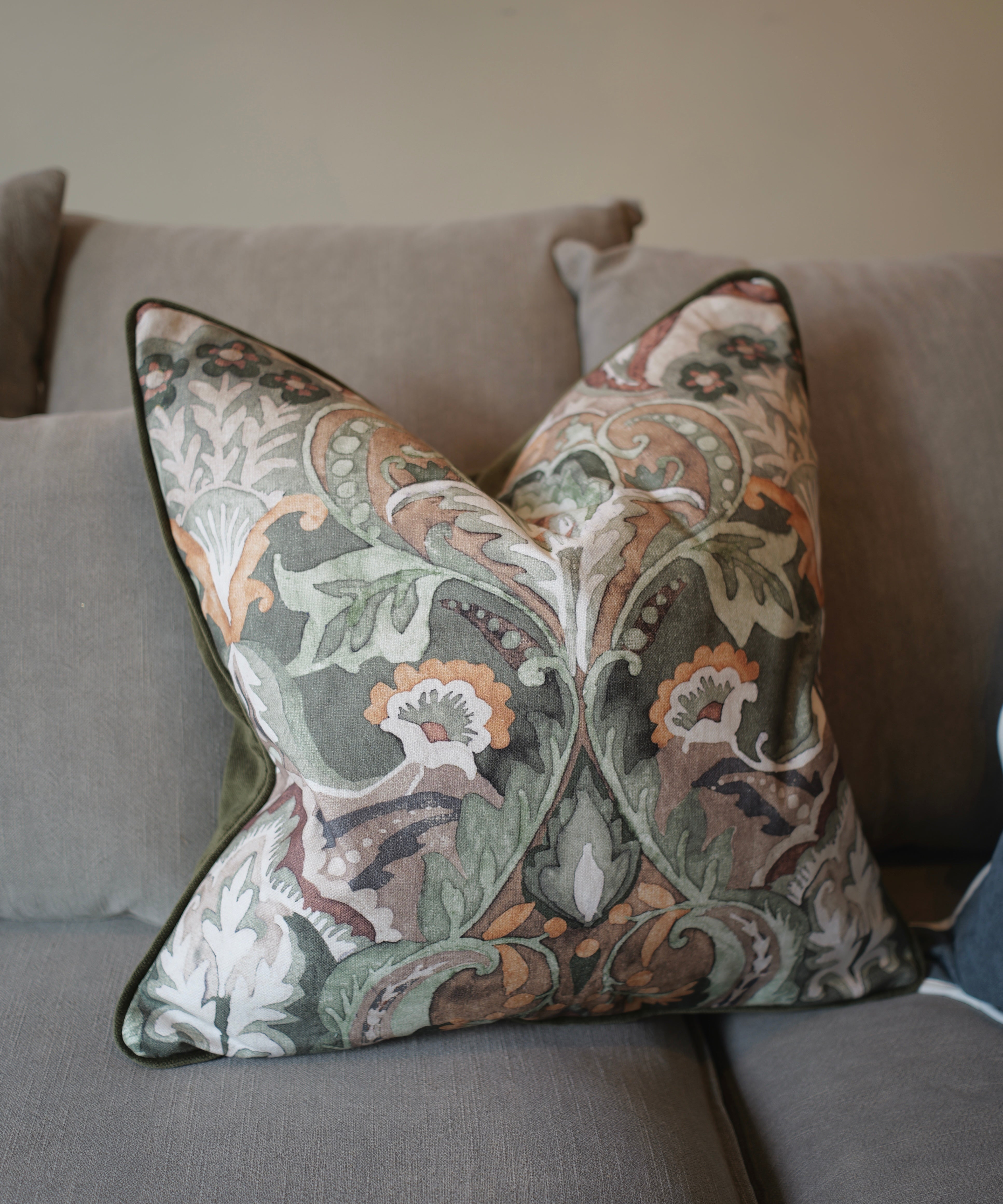A square decorative throw pillow with a detailed botanical and floral damask pattern in muted green, beige, orange, and white, featuring a dark green piped edge, resting on a gray fabric sofa.