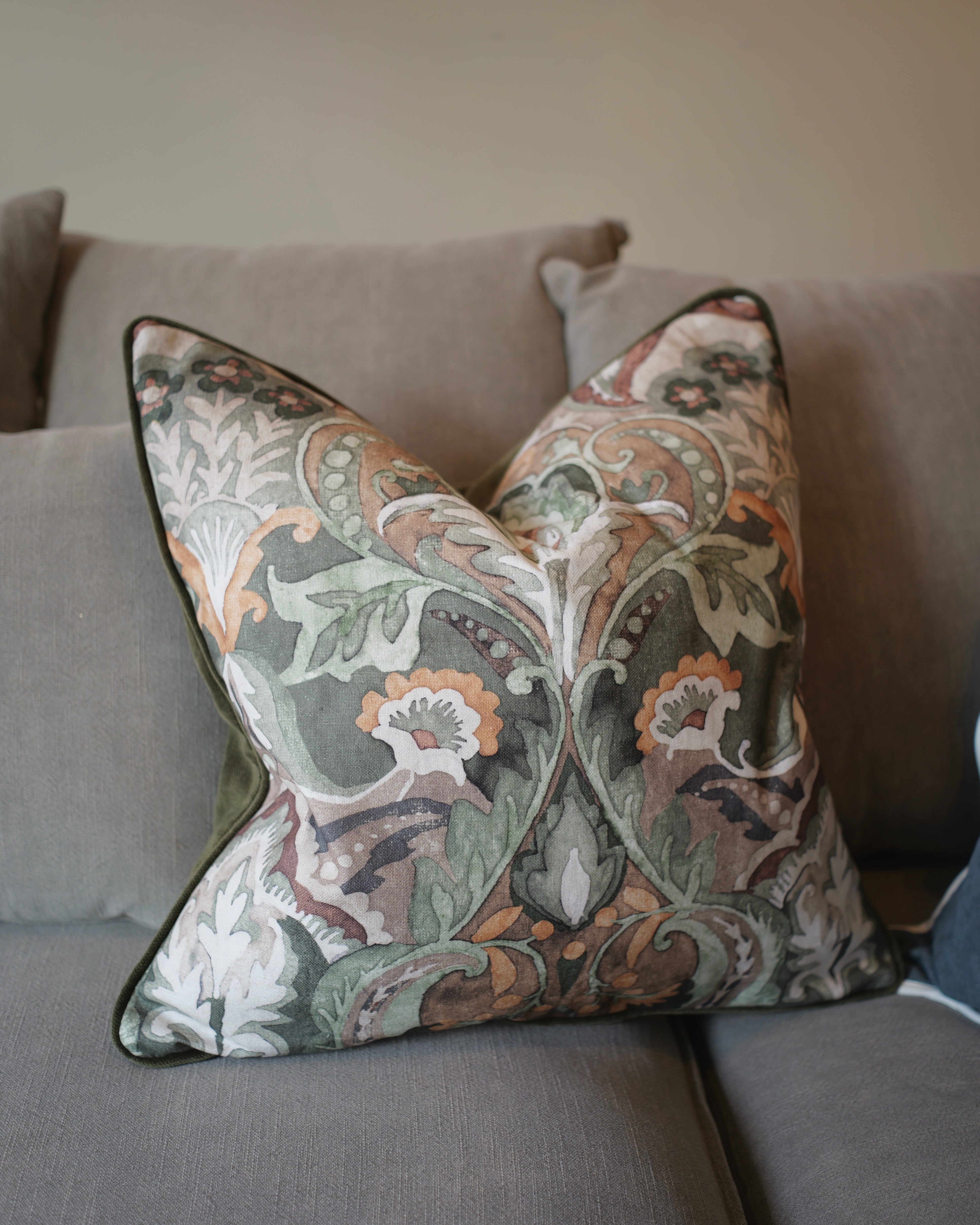 A square decorative throw pillow with a detailed botanical and floral damask pattern in muted green, beige, orange, and white, featuring a dark green piped edge, resting on a gray fabric sofa.