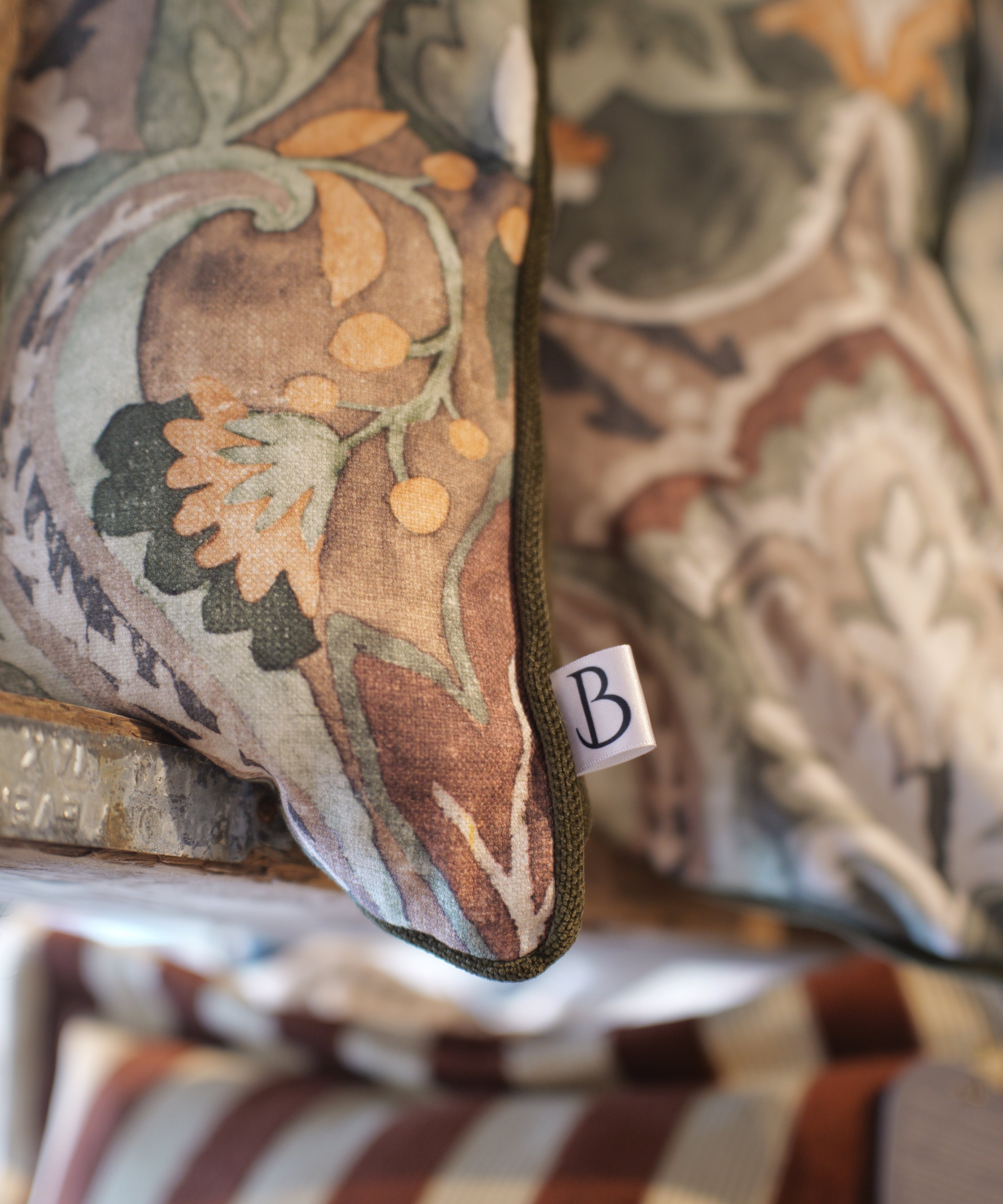 A close-up of a decorative throw pillow with an intricate botanical and floral pattern in muted earthy tones of brown, beige, green, and orange. A small white fabric tag with a black monogram letter 'B' is visible on the side.