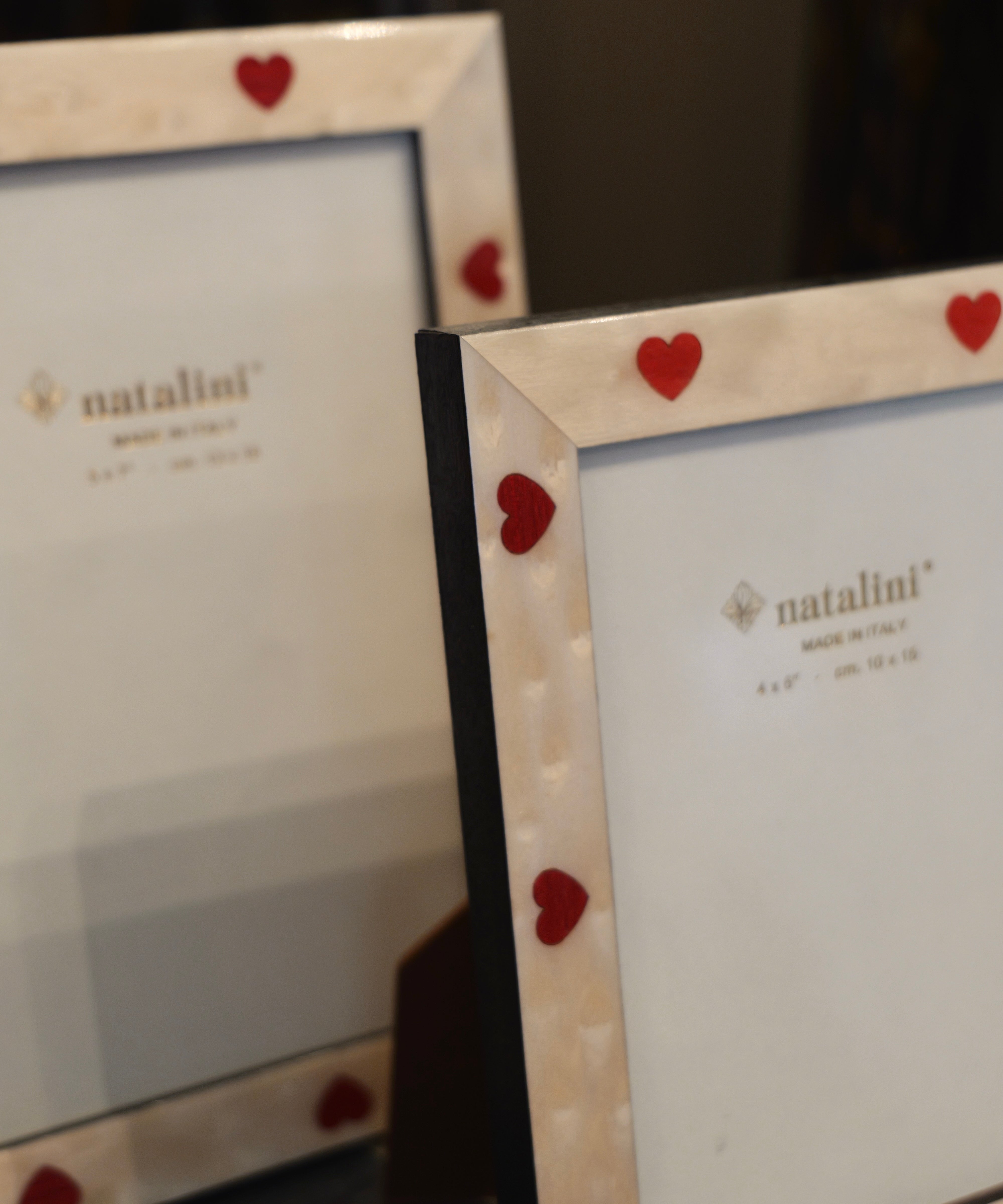 Two white marble-style picture frames with small red heart accents on the corners, shown on a dark surface. The front frame is in focus, displaying the 'natalini' brand name on the insert, while the second frame is softly blurred in the background.