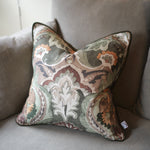 A square decorative throw pillow with an intricate paisley and floral damask pattern in muted tones of green, beige, brown, and orange, resting on a gray fabric sofa.